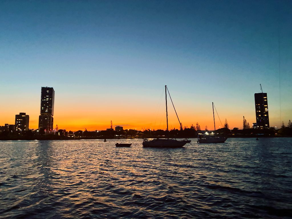 Gold Coast Broadwater at dusk — sailboat silhouettes and city building lights reflecting on the water from a hens party boat